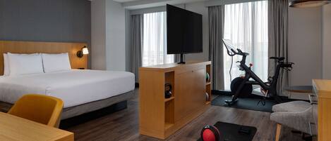 A hotel room with a bed, a desk, a TV, an exercise bike, and a workout mat.