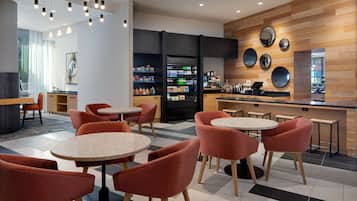 A modern cafe with round tables and orange chairs, a coffee machine, and a refrigerator stocked with beverages.