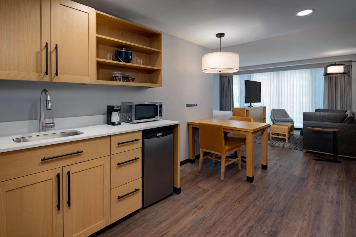 A modern hotel room with a kitchen area, a dining table, and a seating area with a sofa and armchairs.