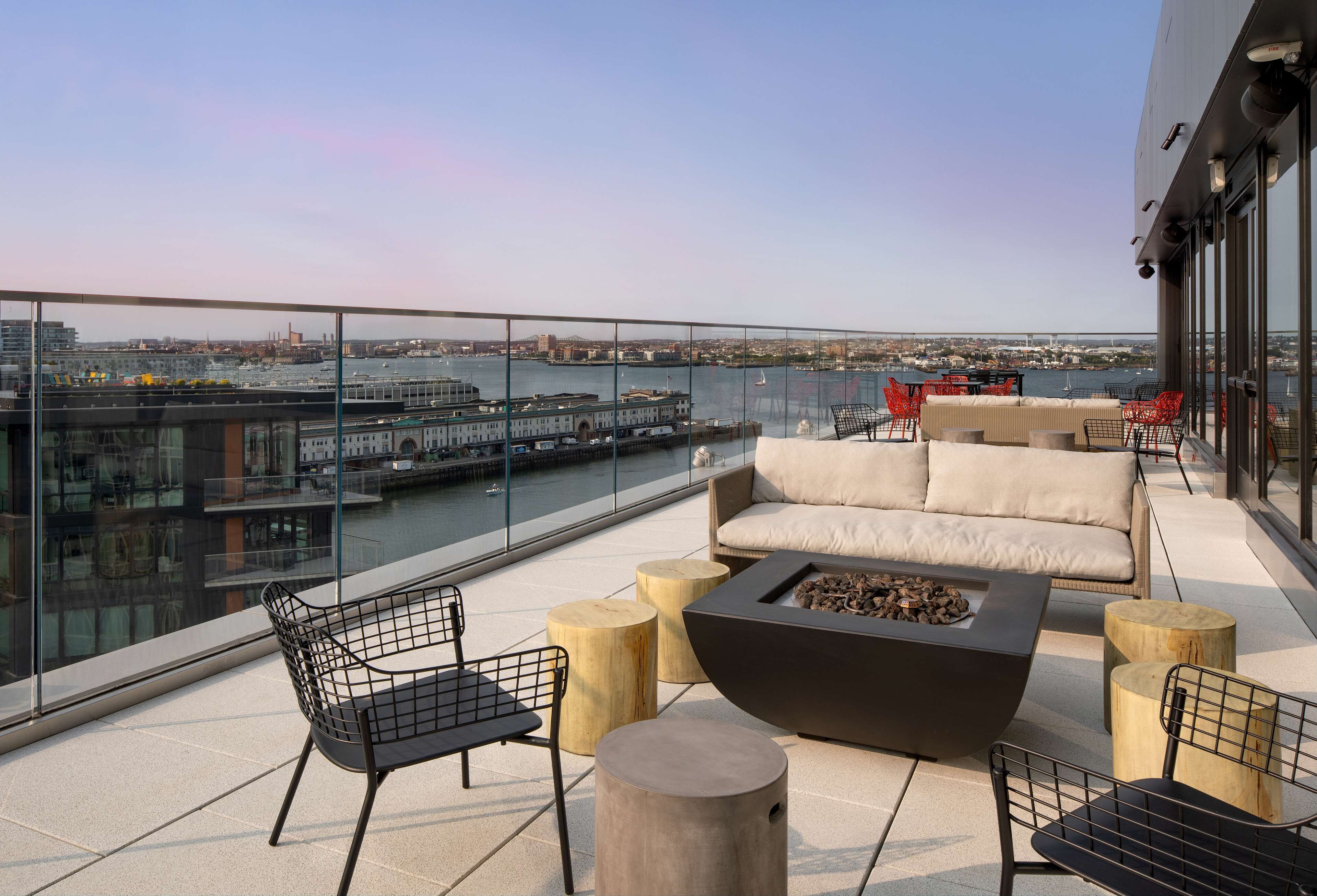 A rooftop terrace with a sofa, chairs, a fire pit, and a view of a cityscape and river.