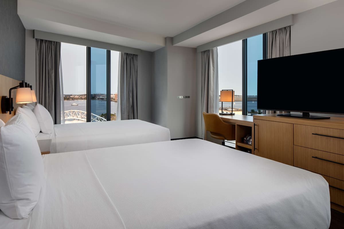 A hotel room with two beds, a TV, a desk, and a view of the sea.
