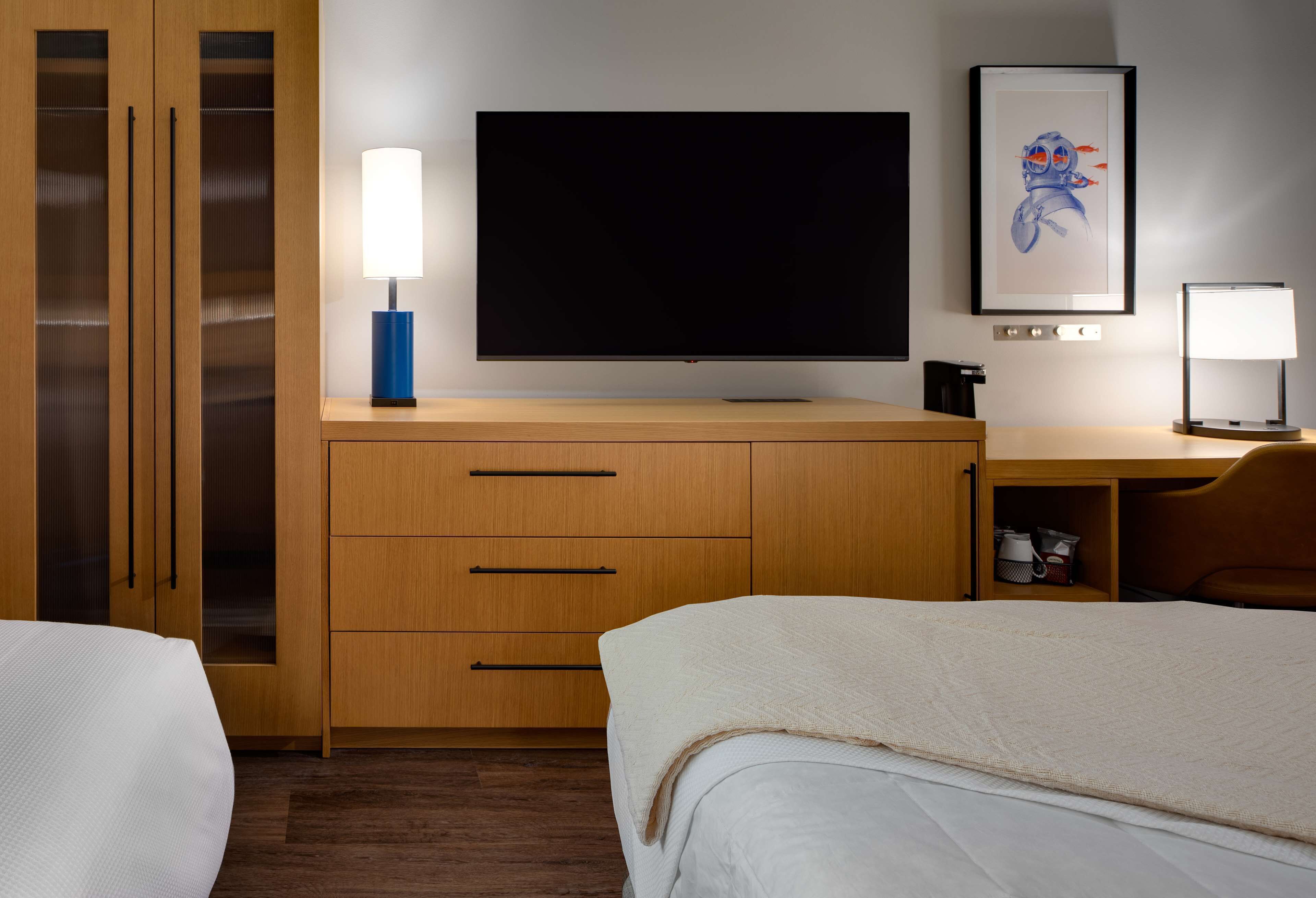 A hotel room with a large flat-screen TV, wooden furniture, a framed artwork, and a desk with a lamp.