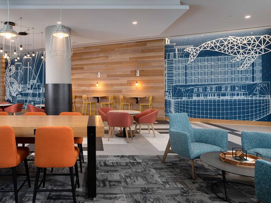 Un moderno vestíbulo de hotel con pared de madera, arte mural geométrico, asientos en tonos naranja y azul, e iluminación colgante.