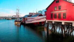 A red building with a sign reading "Boston Tea Party - Ships & Museum" docked by a waterway with a tall ship and a smaller boat. - Hyatt Place Boston/Seaport District (Boston)