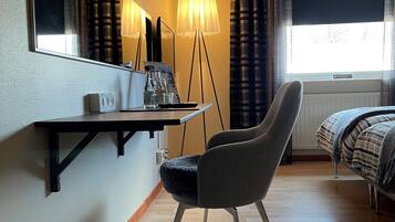 Habitación de hotel con cama, escritorio con lámpara, silla, espejo y ventana con cortinas.