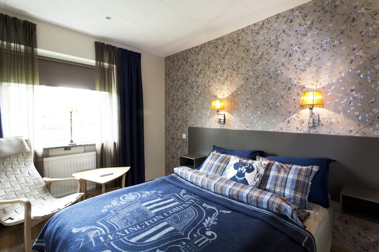 Una habitación de hotel con una cama, una silla, una mesita y dos lámparas fijadas a la pared.