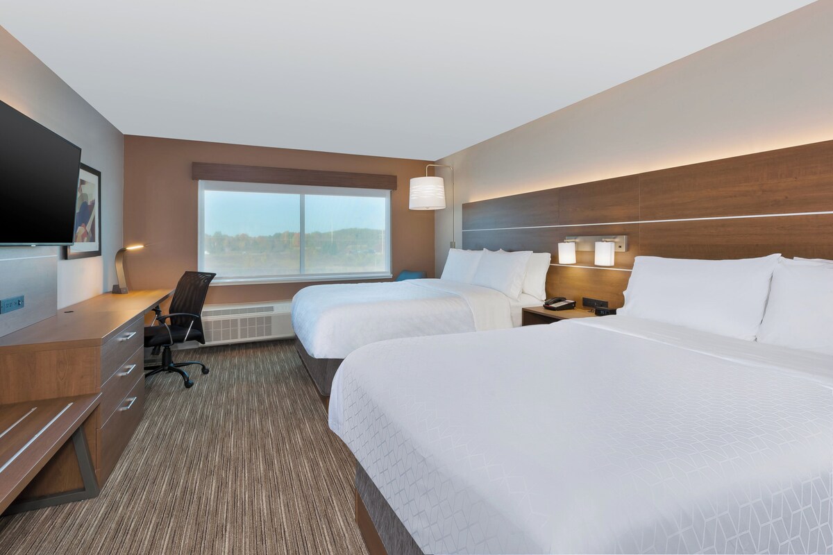 Quarto de hotel com duas camas, uma mesa, uma cadeira, uma TV e uma janela grande.