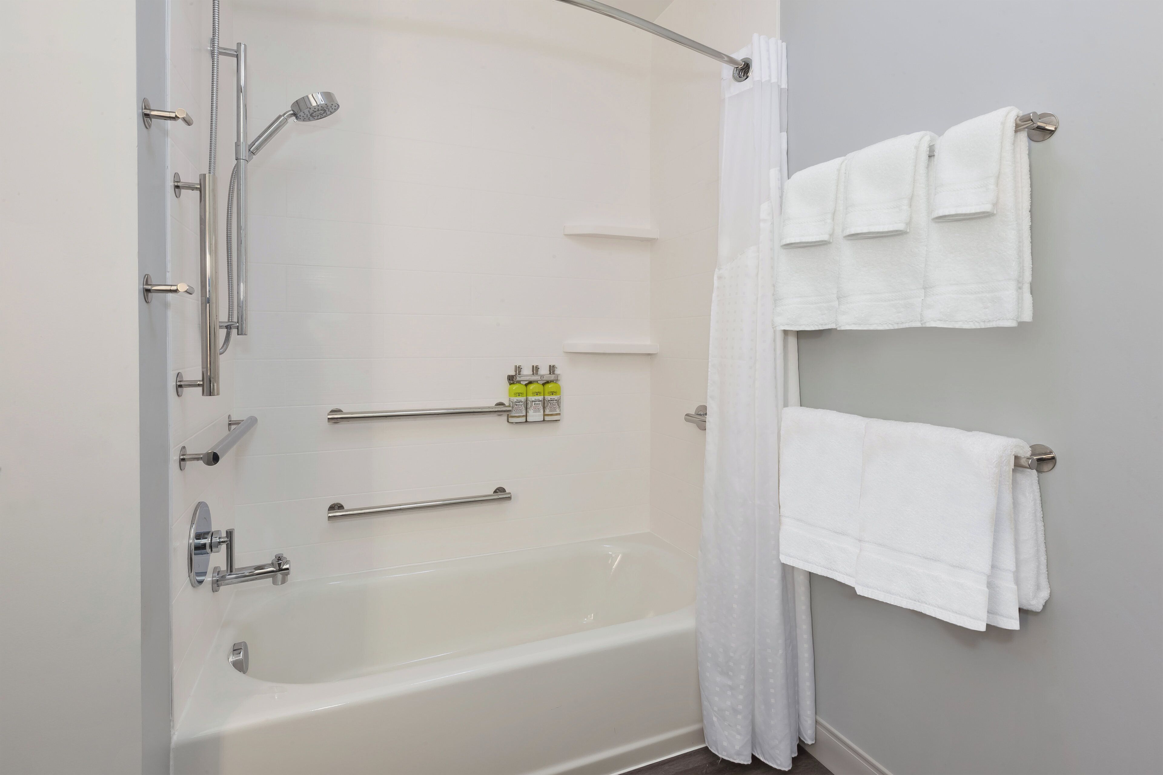 Une salle de bain équipée d’une baignoire, d’une douche et de barres d’appui. On y trouve des serviettes sur un porte-serviettes et un rideau de douche.