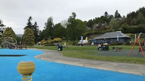 Een speeltuin met een blauw oppervlak, een geel en grijs zandspeeltoestel, en een gebouw met een blauw dak op de achtergrond.