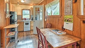 Interior de una cabaña con paredes de madera, una mesa de comedor, sillas, una cocina con estufa, refrigerador y una ventana con vistas a la vegetación.