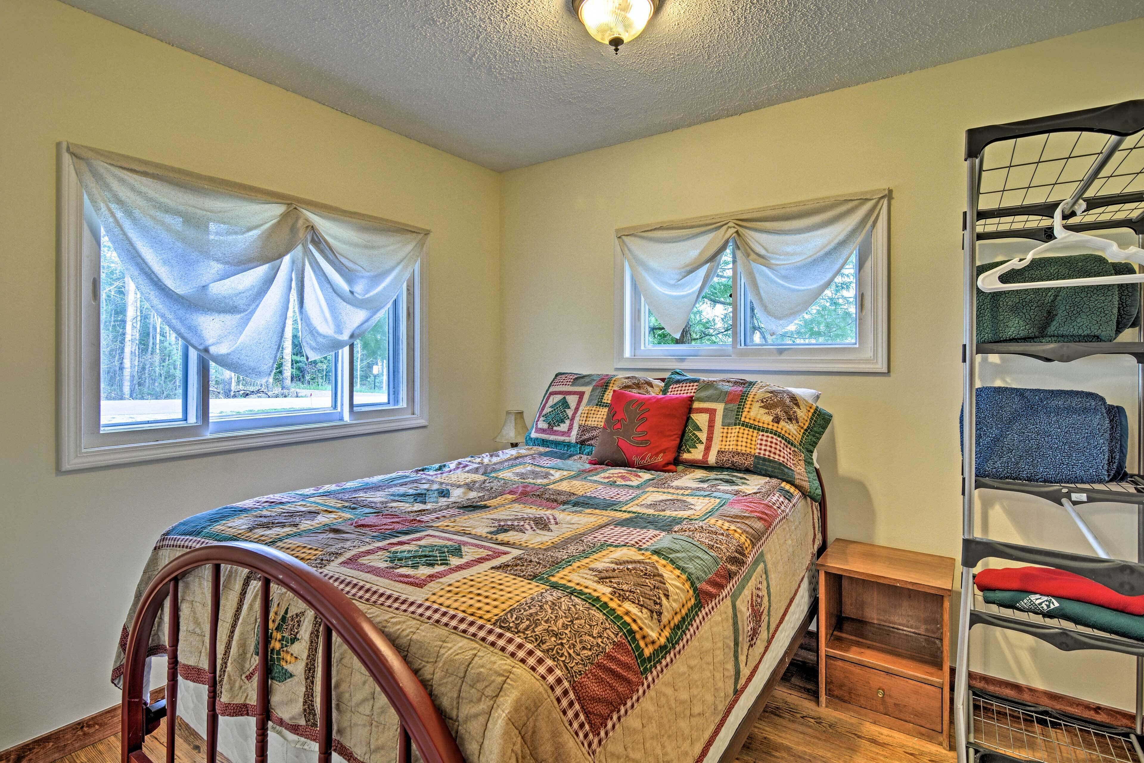 A bedroom with a wooden bed, a quilt, two windows with curtains, a nightstand, and a metal rack with clothes.