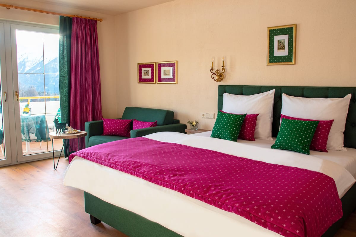 Habitación de hotel con una cama grande, un sofá verde y una pequeña mesa. Hay dos cuadros en la pared y una ventana con cortina.