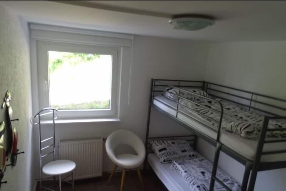 A dormitory room with bunk beds, a radiator, a chair, and a window with a view of greenery.