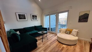 A living room with a dark green sectional sofa, a round beige sofa, a small coffee table, and a large sliding glass door leading to a balcony.
