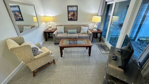 A living room with a sofa, armchair, coffee table, and a TV. There are two lamps on side tables, and a large mirror on the wall.