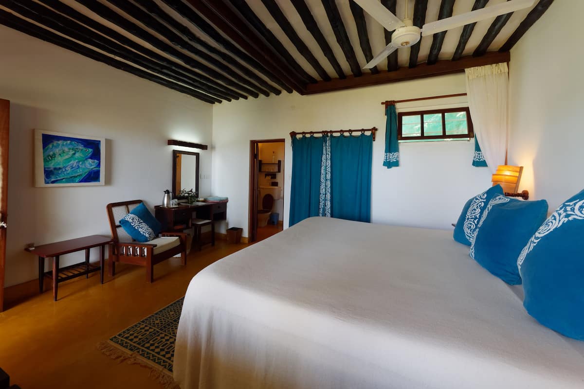 A bedroom with a large bed, a desk with a chair, a ceiling fan, and a painting on the wall.