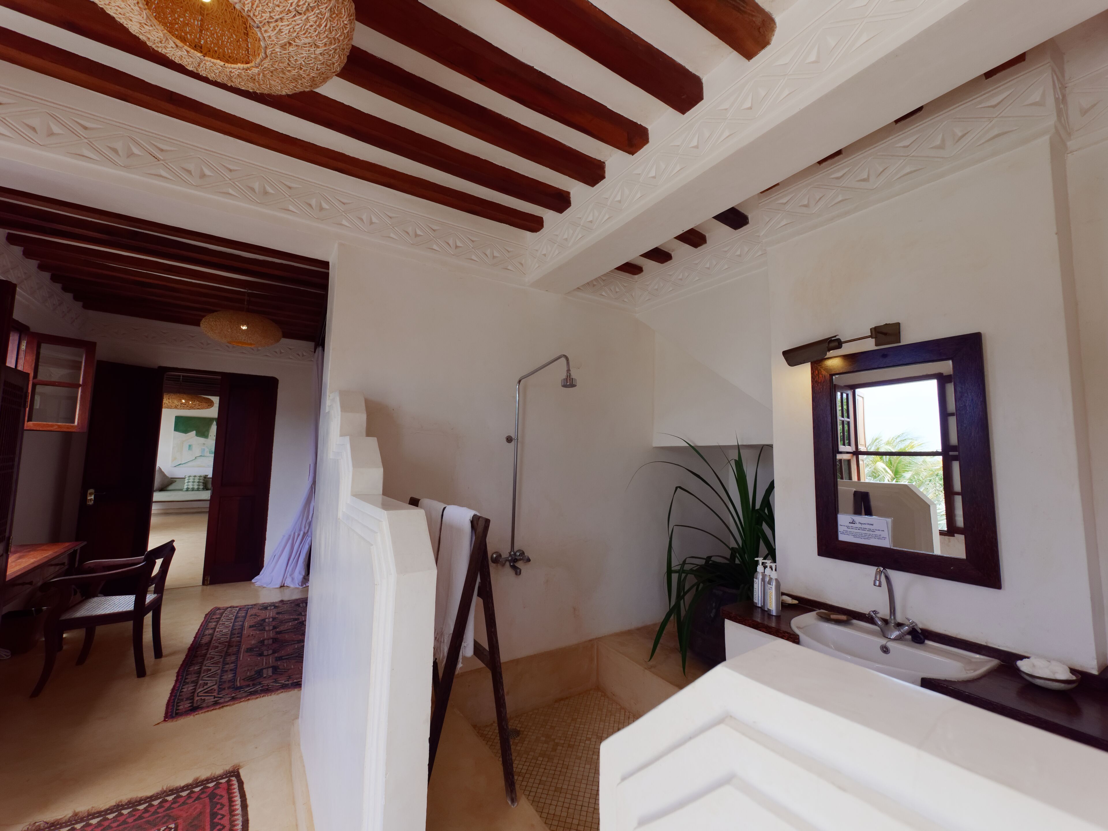 A bathroom with a shower, sink, and a large mirror. There is a wooden ceiling and a patterned rug on the floor.