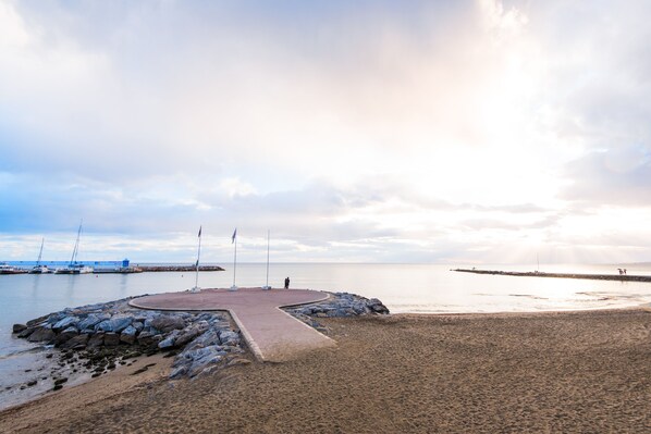 Una platja amb passeig pavimentat, línia de costa rocosa i velers a l'aigua.