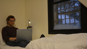 Eine Person sitzt auf einem Bett, auf dem ein Laptop steht, mit einem Fenster und Blick auf Bäume sowie einer Wandsteckdose.