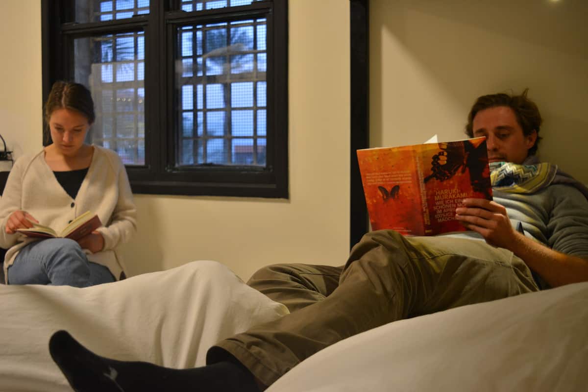 Två personer sitter på en säng och läser böcker i ett rum med ett fönster och en enkel vägg.