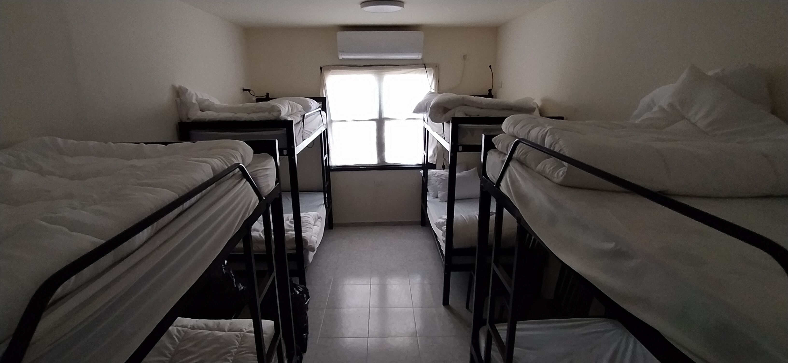 A dormitory room with bunk beds arranged in two rows, a window with blinds, and a ceiling fan.