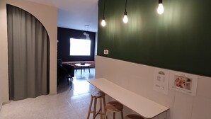 Ein modernes Interieur mit grüner Akzentwand, einem weißen Tisch und zwei Hockern. Ein Fenster mit Aussicht, eine Tür mit Vorhang und eine Wandleuchte sind ebenfalls zu sehen.