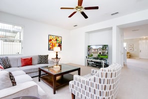 A living room with a sofa, coffee table, and patterned chairs. A ceiling fan is visible, and a television is mounted on the wall.