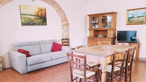 A dining area with a wooden table and chairs, a sofa with red cushions, a cabinet with dishes, and a painting on the wall. - Agriturismo Il Poderino (Bibbona)