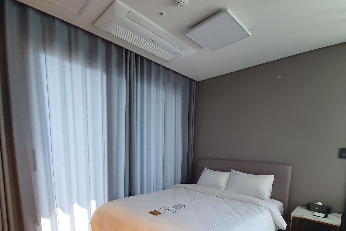 A neatly made bed with white linens, a wooden floor, and a nightstand with a lamp and a trash bin.