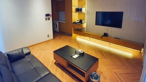 Un salon moderne avec un canapé noir, une table basse en bois, un téléviseur fixé au mur et un coin cuisine équipé d’un four à micro-ondes et d’un lave-linge.