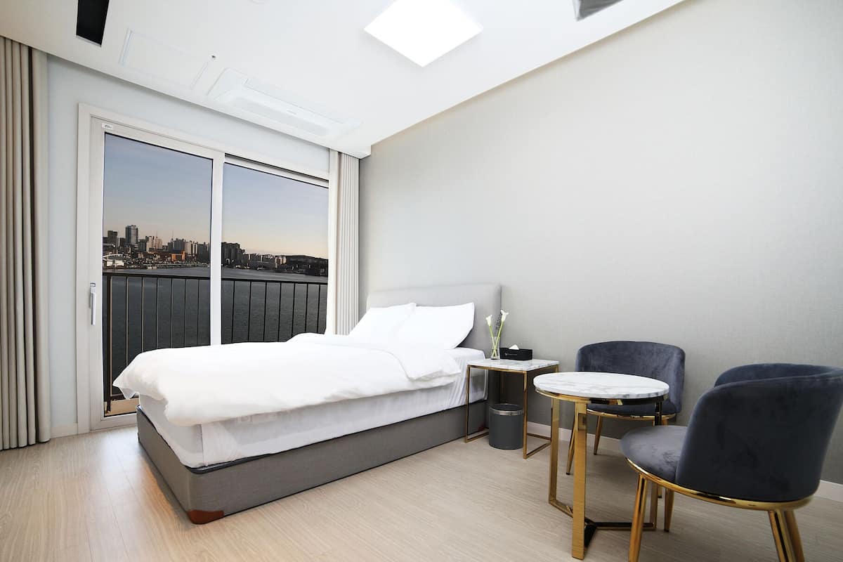A hotel room with a bed, a small table, two chairs, a nightstand, and a view of a city skyline through a large window.