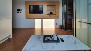 Un salon moderne doté d’un grand téléviseur à écran plat fixé sur un panneau en bois, d’une table basse en marbre avec une télécommande, et d’une petite étagère sur laquelle se trouve un téléphone.
