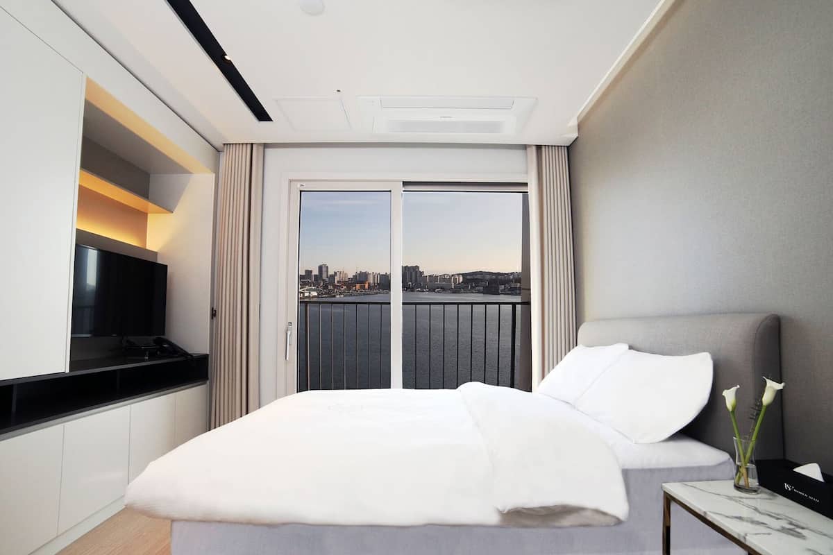 A hotel room with a bed, a TV, a bedside table with a vase, and a view of a cityscape through a large window.