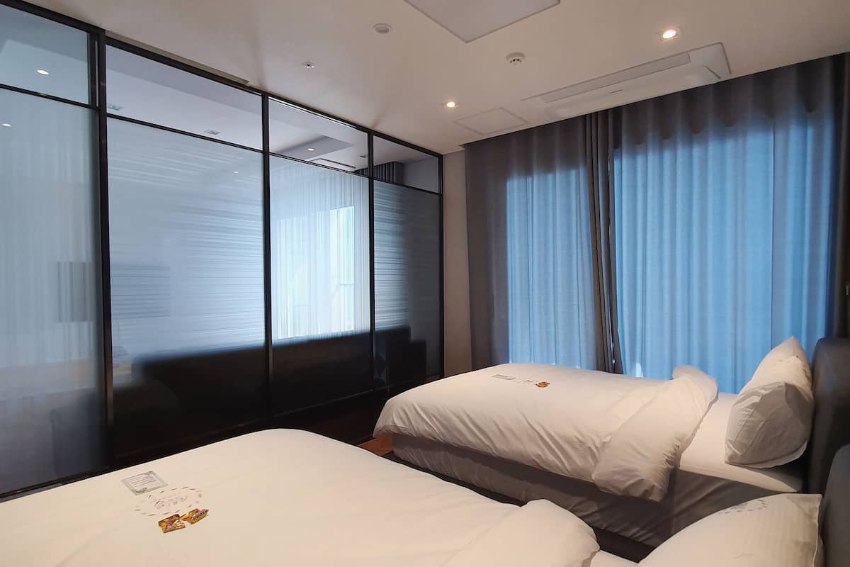 A hotel room with two beds, a large window with blinds, and a ceiling light.