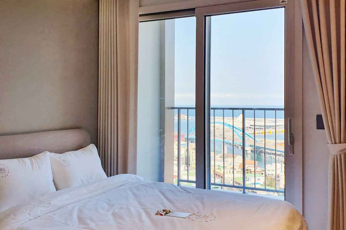 A hotel room with a large bed, a view of a cityscape through a sliding glass door, and a ceiling with an air conditioning unit.