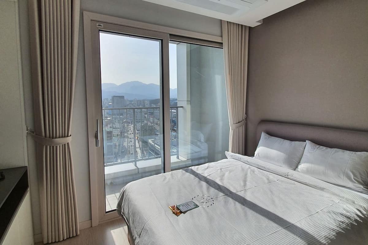 A modern hotel room with a large bed, a view of a cityscape through floor-to-ceiling windows, and a ceiling with recessed lighting.