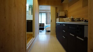 Un couloir étroit mène à une chambre où se trouvent un lit et une fenêtre. On aperçoit, sur la droite, une petite cuisine équipée d’une cuisinière et de placards.