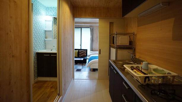 Une cuisine compacte équipée d’un évier, d’une cuisinière et d’un égouttoir. Un petit coin repas avec une table et des chaises est visible à travers une porte ouverte.