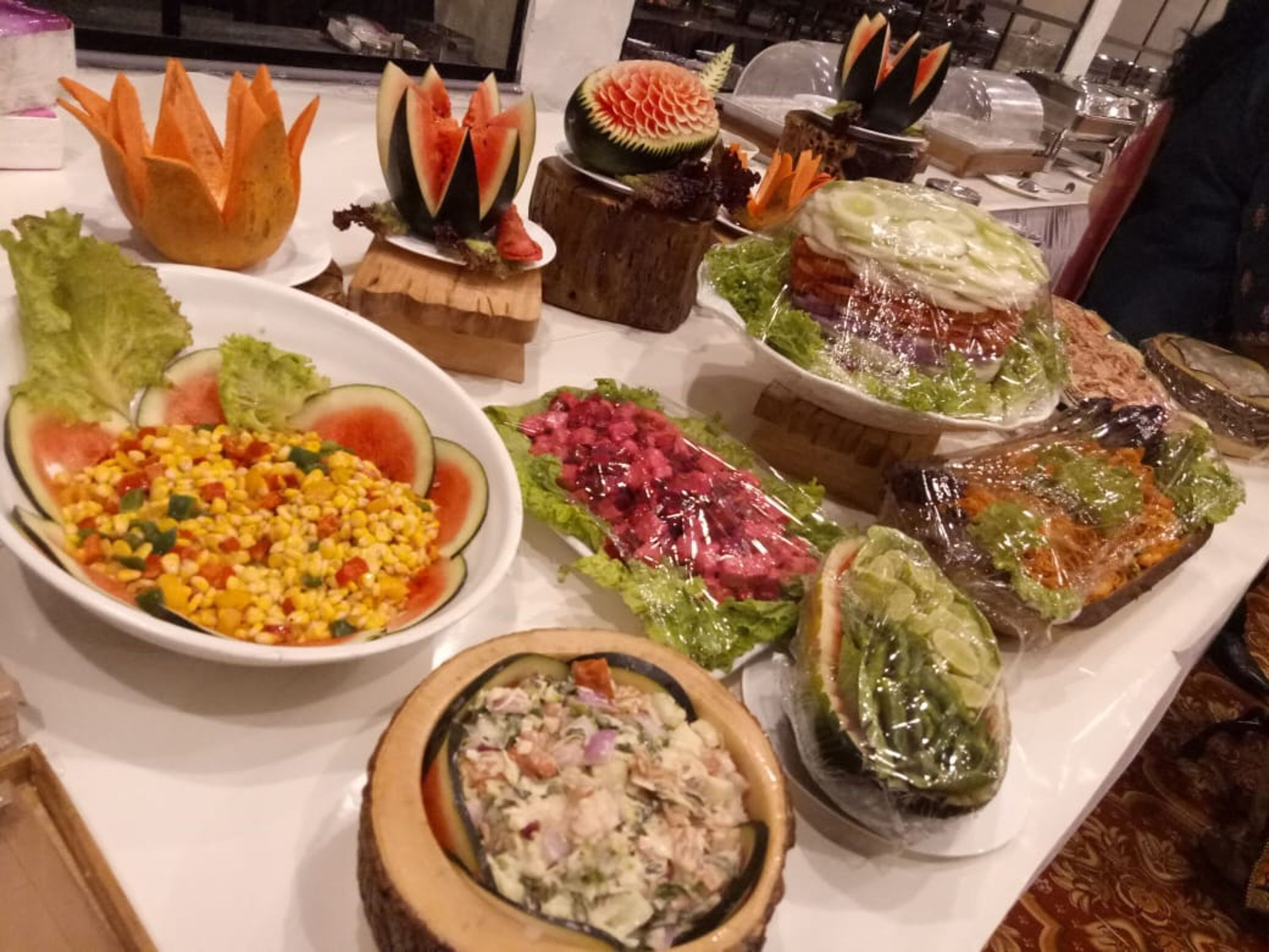 A variety of food items displayed on a table, including salads, a bowl of corn salad, and creatively carved fruits.