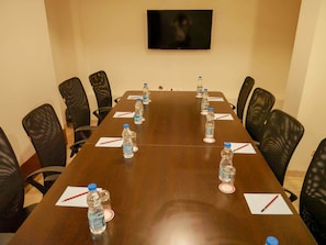 A conference room with a long table, black mesh chairs, water bottles, and red pens on napkins. - Emporio Hotel (Haldwani)
