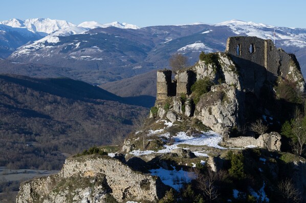 A historic stone ruin with a flag on top, surrounded by snow-covered mountains and a forested valley.