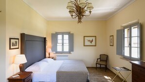 Un dormitorio con cama, lámpara de noche, cómoda, silla y una ventana con contraventanas.