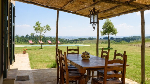 Una mesa y sillas de madera bajo un techo de paja, con vistas a una zona cubierta de césped, árboles y una lejana vista de colinas.