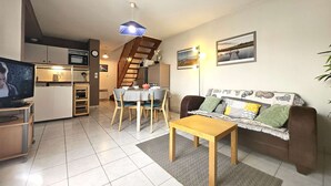 Un salon comprenant un canapé, une table basse, une table à manger et des chaises. On y trouve également un coin cuisine avec un four à micro-ondes, un four et un évier. Un escalier est visible au fond.