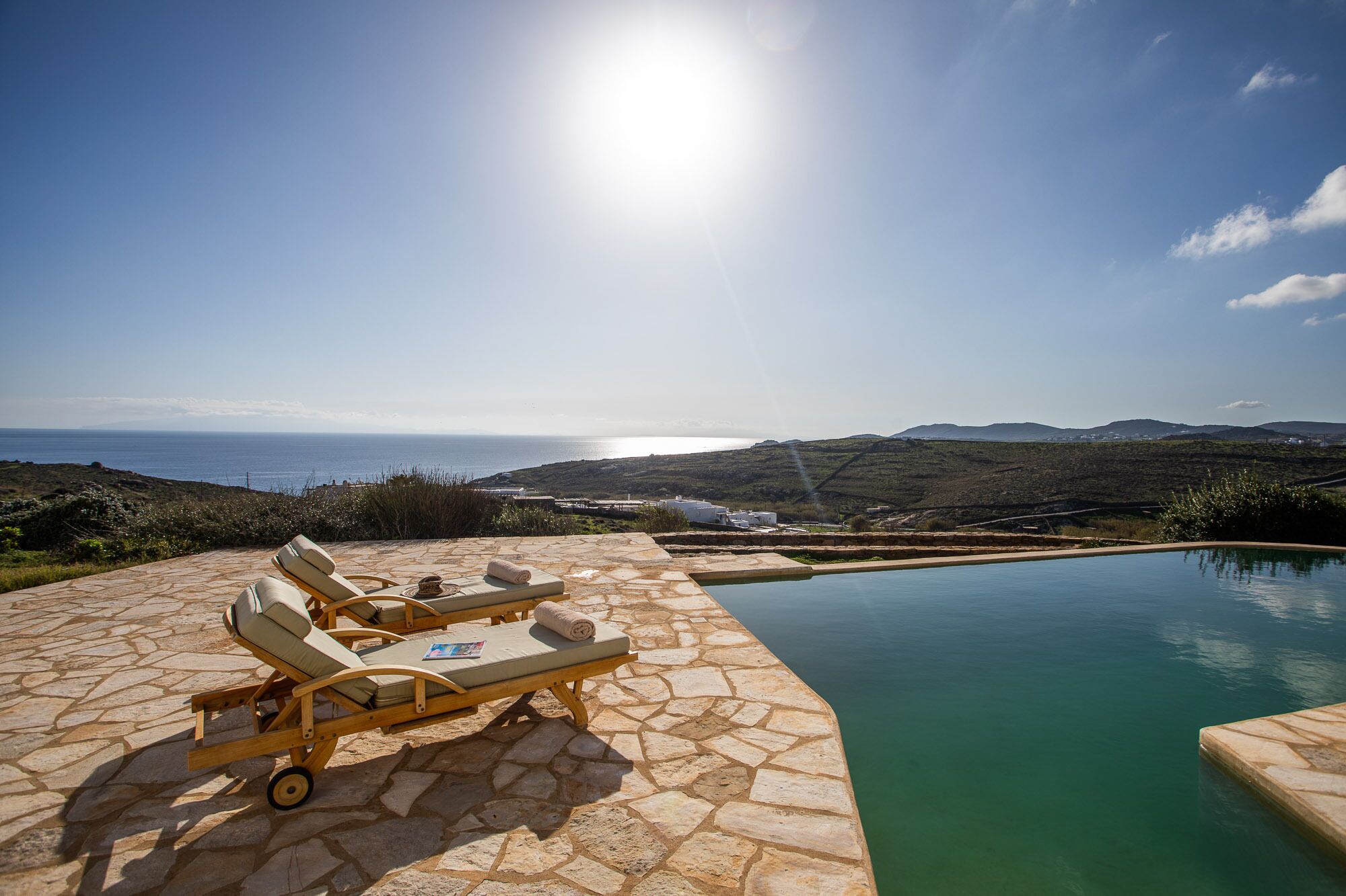 En stenbelagt terrasse med liggestole og en swimmingpool med udsigt over et kystlandskab.