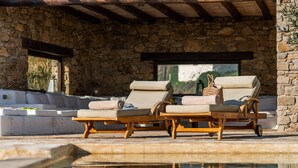 A poolside area with two wooden lounge chairs, a stone wall, and a view of the outdoors. - Zen Residence in Lia beach (Mykonos)
