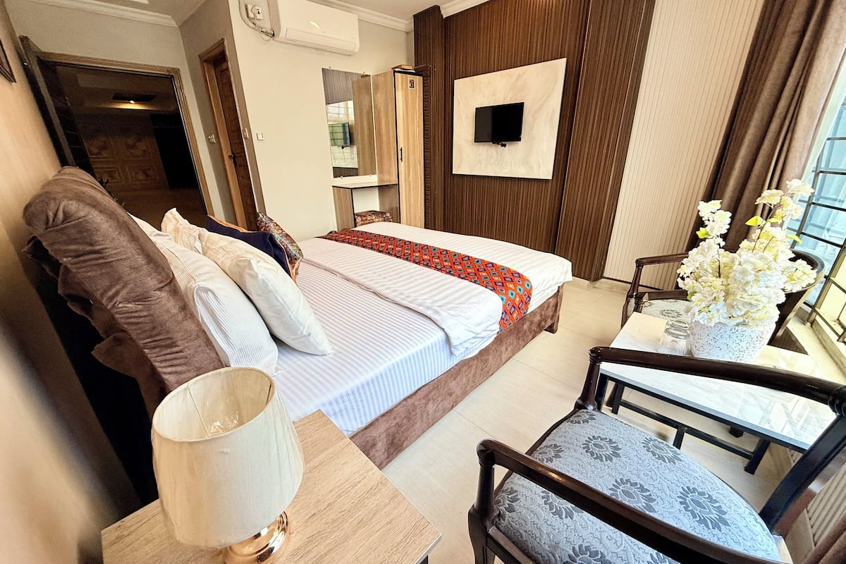 A hotel room with a bed, a TV mounted on the wall, a table with a lamp, and a chair.