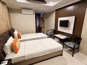 A hotel room with two beds, a TV mounted on the wall, a small table with chairs, and an air conditioning unit.
