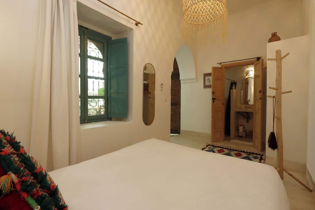 A bedroom with a bed, a window with green shutters, a mirror, and a wardrobe.