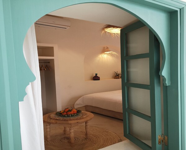 Una habitación con una cama, una mesita con fruta, una lámpara de pared y una puerta con paneles de vidrio esmerilado.